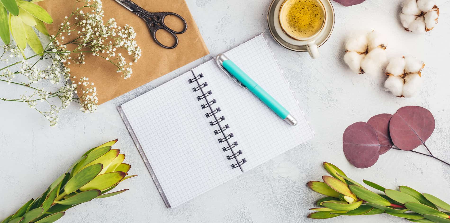 Ein Notizblock und Stift, umgeben von Pflanzen und Blüten, neben einer Tasse Kaffee, als Symbol für die sorgfältige Planung eines Gartens